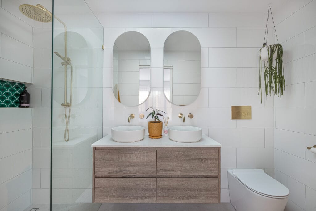 Modern bathroom with double vanity, oval mirrors, gold fixtures, glass shower, hanging plant, and white tiled walls