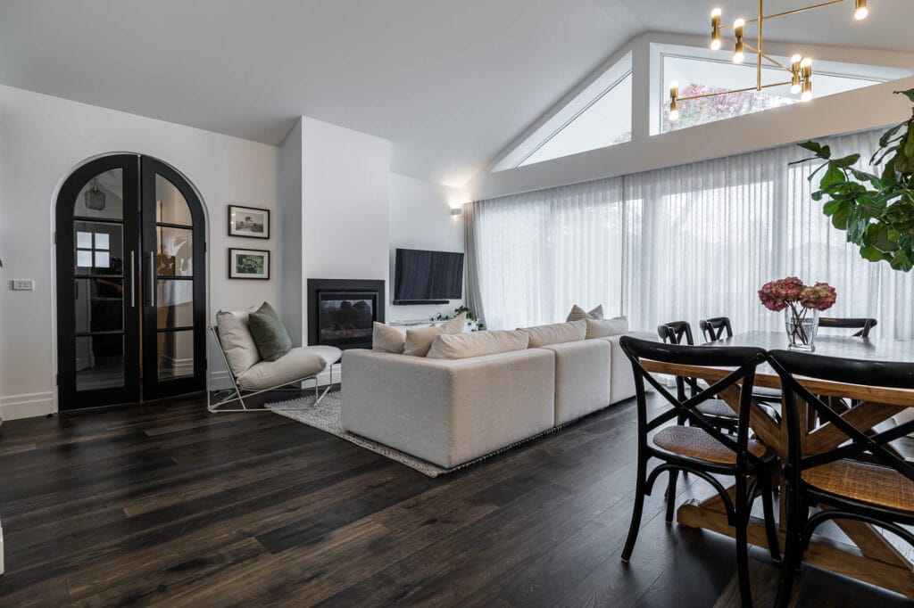 Modern living and dining area with dark wood flooring, white sofa, arched black doors, large windows, and soft natural light
