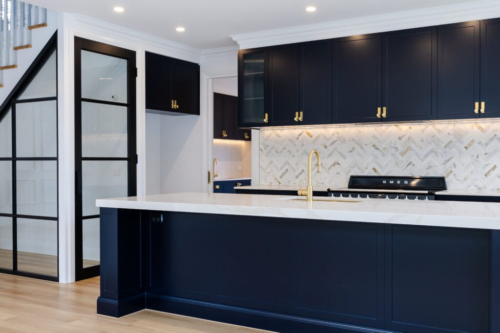 Modern kitchen with navy cabinets, gold hardware, white marble countertops, herringbone backsplash, and black glass doors